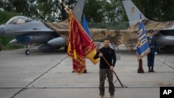 Президент України Володимир Зеленський з прапором військової частини з нагоди Дня Повітряних Сил на фоні винищувачів F-16 у невстановленому місці в Україні, неділя, 4 серпня 2024 року. AP Photo/Efrem Lukatsky