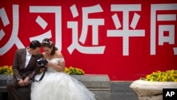 资料照:一对穿着婚礼服装的夫妇2018年5月19日在北京观看照相机背面的照片,身后是关于中国国家主席习近平的宣传口号。