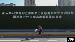一名戴着口罩的妇女从武汉街头一幅宣传习近平湖北讲话的大标语前骑车而过。(2020年3月18日)